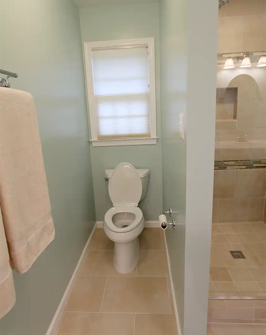 Bristow VA primary bathroom remodel with private toilet area, soft blue-green walls, beige floor tile, and nearby glass tile shower
