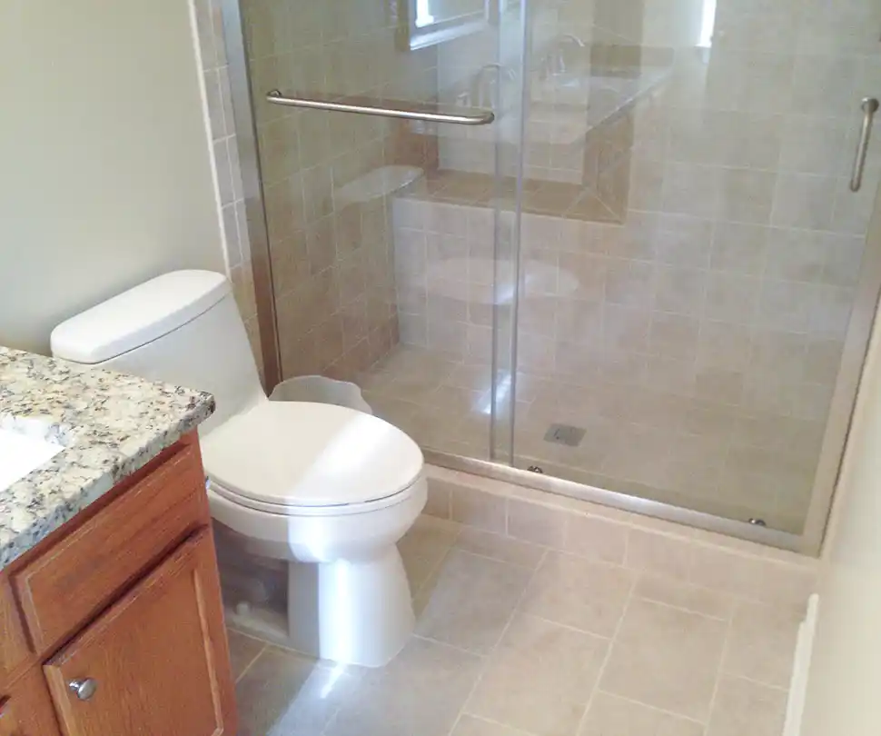 Gainesville VA bathroom remodel with glass-enclosed tile shower, wood vanity, granite countertop, toilet, and neutral floor tile