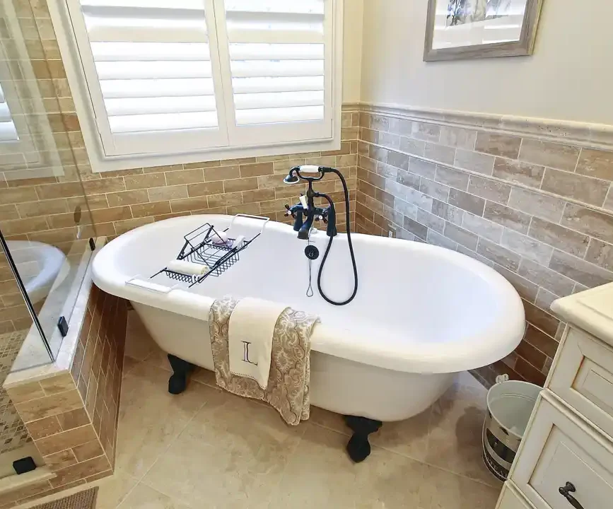 Gainesville VA primary bathroom remodel with clawfoot tub, black tub filler, tile wainscoting, plantation shutters, and glass shower