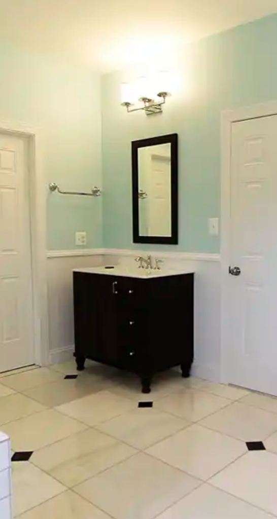 Manassas VA primary bathroom remodel with separate dark vanities, glass subway tile shower, beadboard wainscoting, and marble-look floor tile