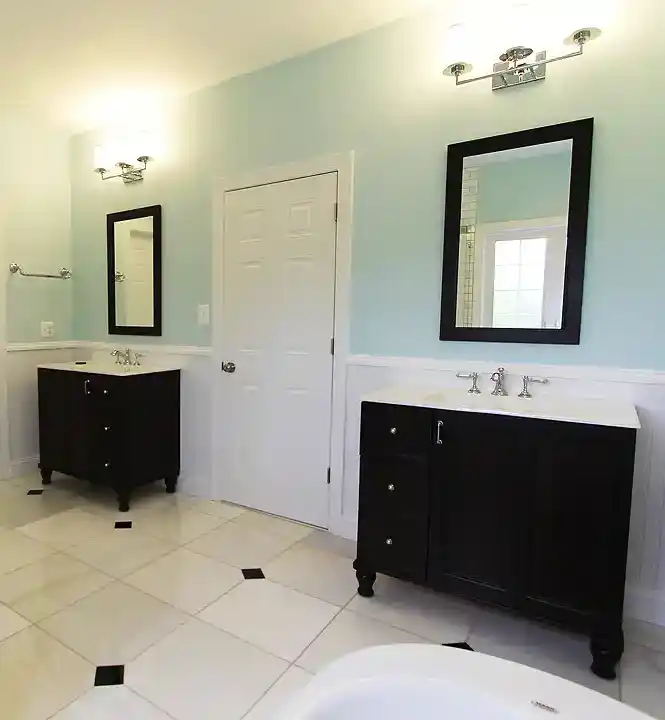 Manassas VA primary bathroom remodel with separate dark vanities, glass subway tile shower, beadboard wainscoting, and marble-look floor tile