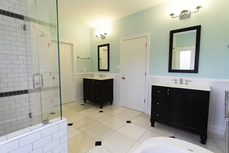Manassas VA primary bathroom remodel with separate dark vanities, glass subway tile shower, beadboard wainscoting, and marble-look floor tile